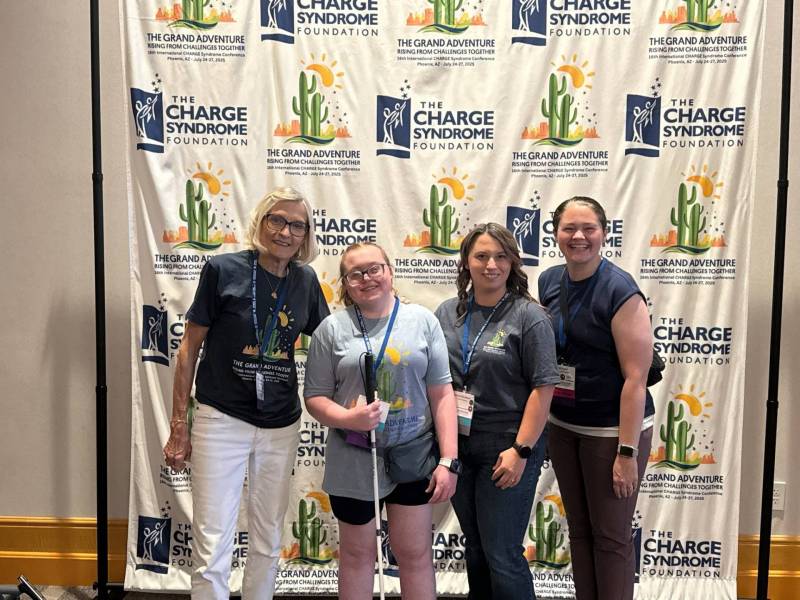 Lanna, Ryder, Kaylee, and Lacey stand in front of a backdrop that has cacti on it as well as text that reads "the CHARGE Syndrome Foundation"